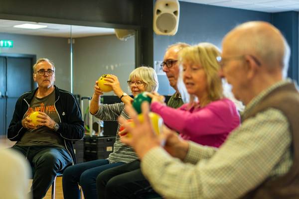 ine Gruppe älterer Menschen sitzt im Halbkreis auf Stühlen in einem hellen Raum und führt gemeinsam Bewegungsübungen mit bunten Gymnastikbällen durch. Alle wirken konzentriert und motiviert, einige lächeln dabei. Die Atmosphäre ist aktiv und gemeinschaftlich.